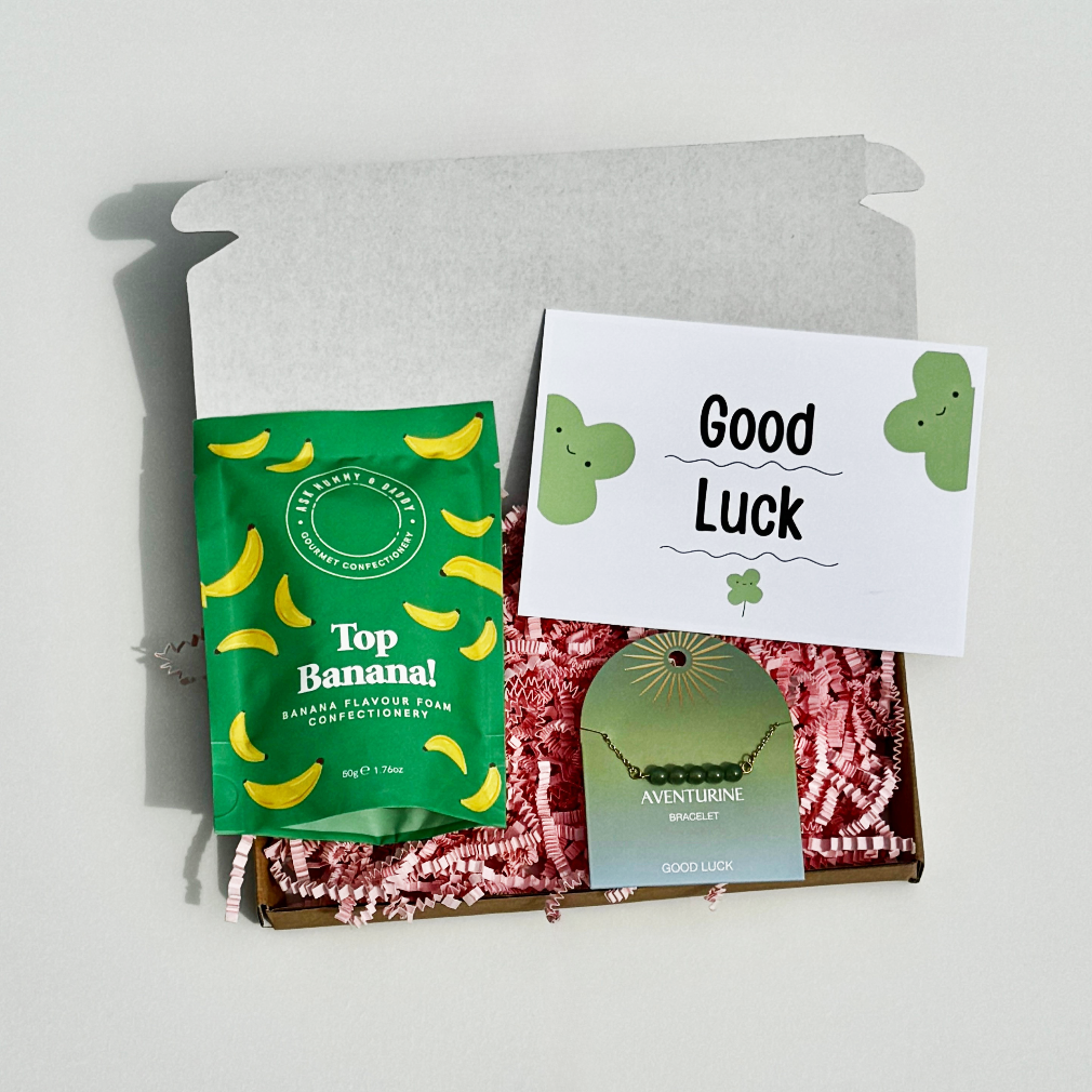 Mini good luck gift box with “Top Banana” banana foam sweets, aventurine crystal bracelet, and a good luck card on pink shredded tissue.