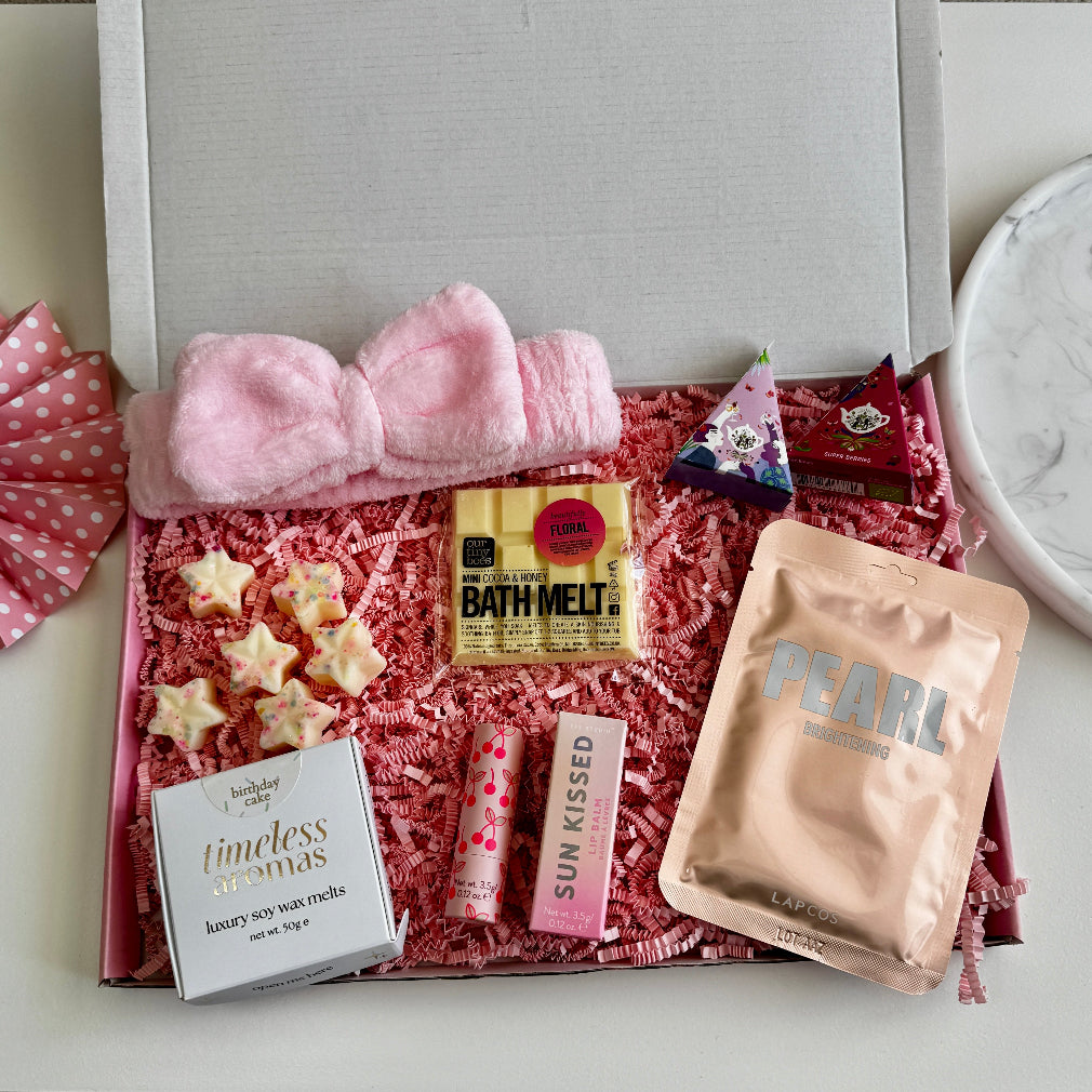 Flat lay of a pink-themed pamper gift box featuring a pink bow headband, PEARL face mask, birthday cake soy wax melts, floral bath melt, lip balms, tea pyramids, and white chocolate star treats.