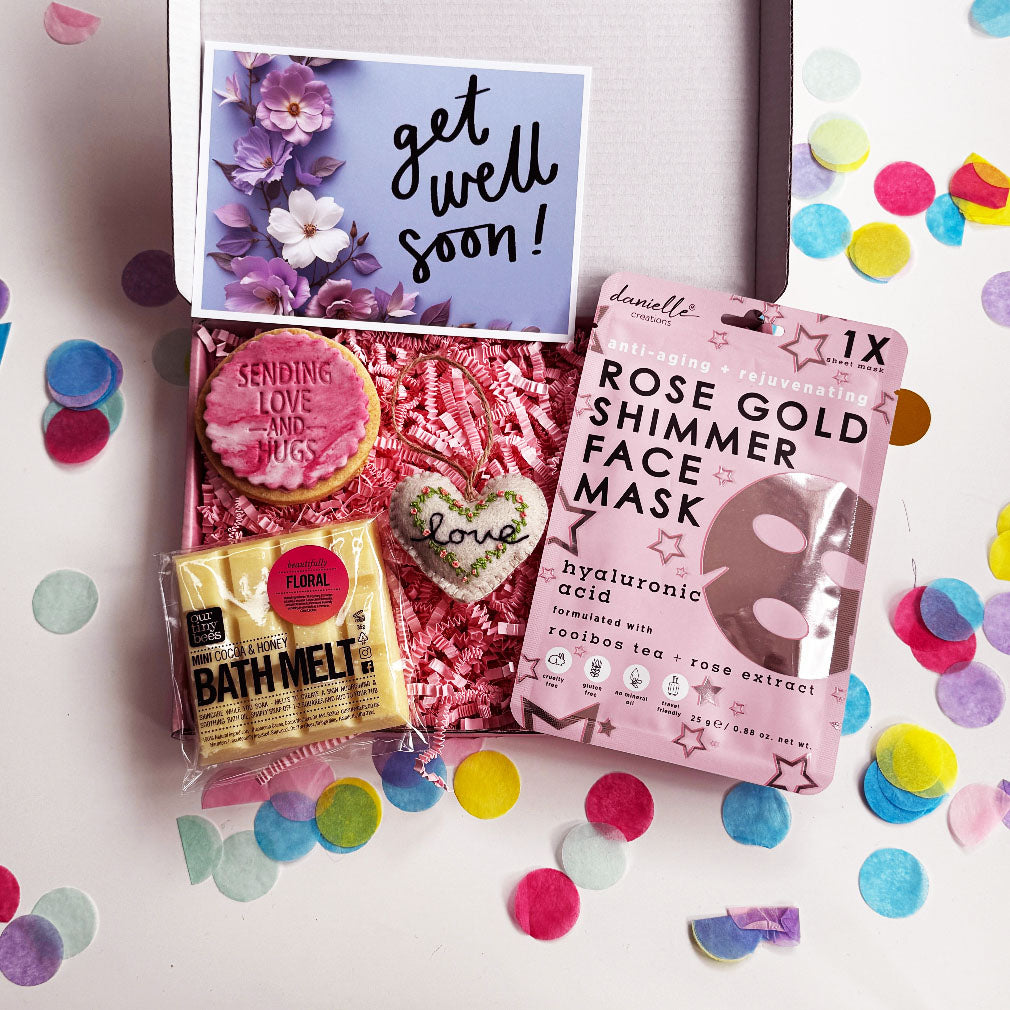 Get well soon gift box including a pink shimmer face mask, floral bath melt, “Sending Love and Hugs” iced biscuit, embroidered heart ornament, and a floral “Get Well Soon” card, surrounded by colourful confetti.