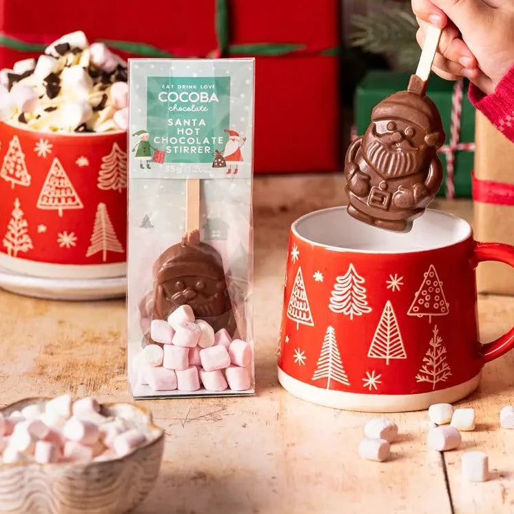 Person stirring a hot chocolate mug with a Santa-shaped chocolate stirrer, surrounded by Christmas decorations and gifts.