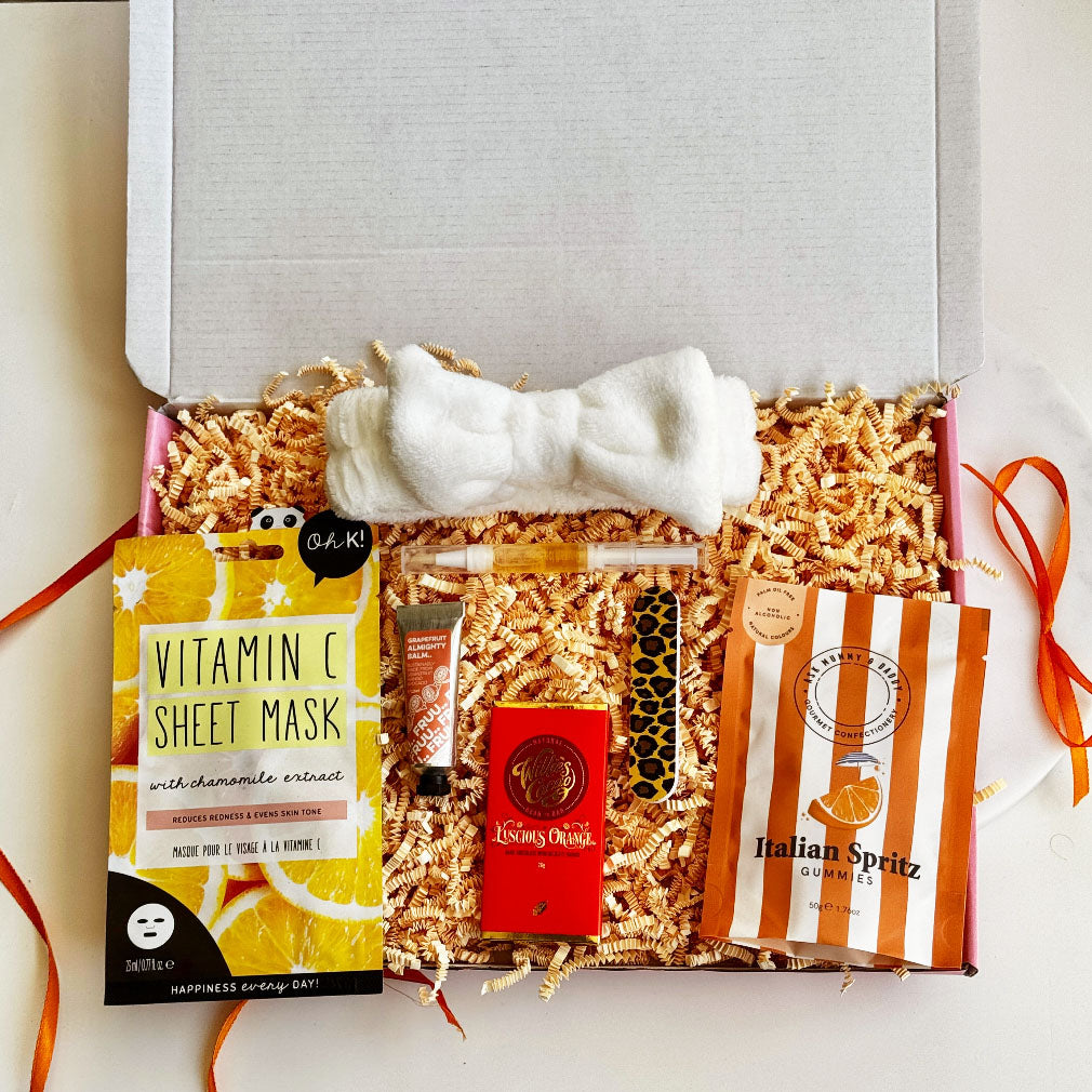Self-care gift box with Vitamin C face mask, Italian Spritz gummies, orange chocolate bar, lip balm, white headband, perfume pen, and leopard print nail file.
