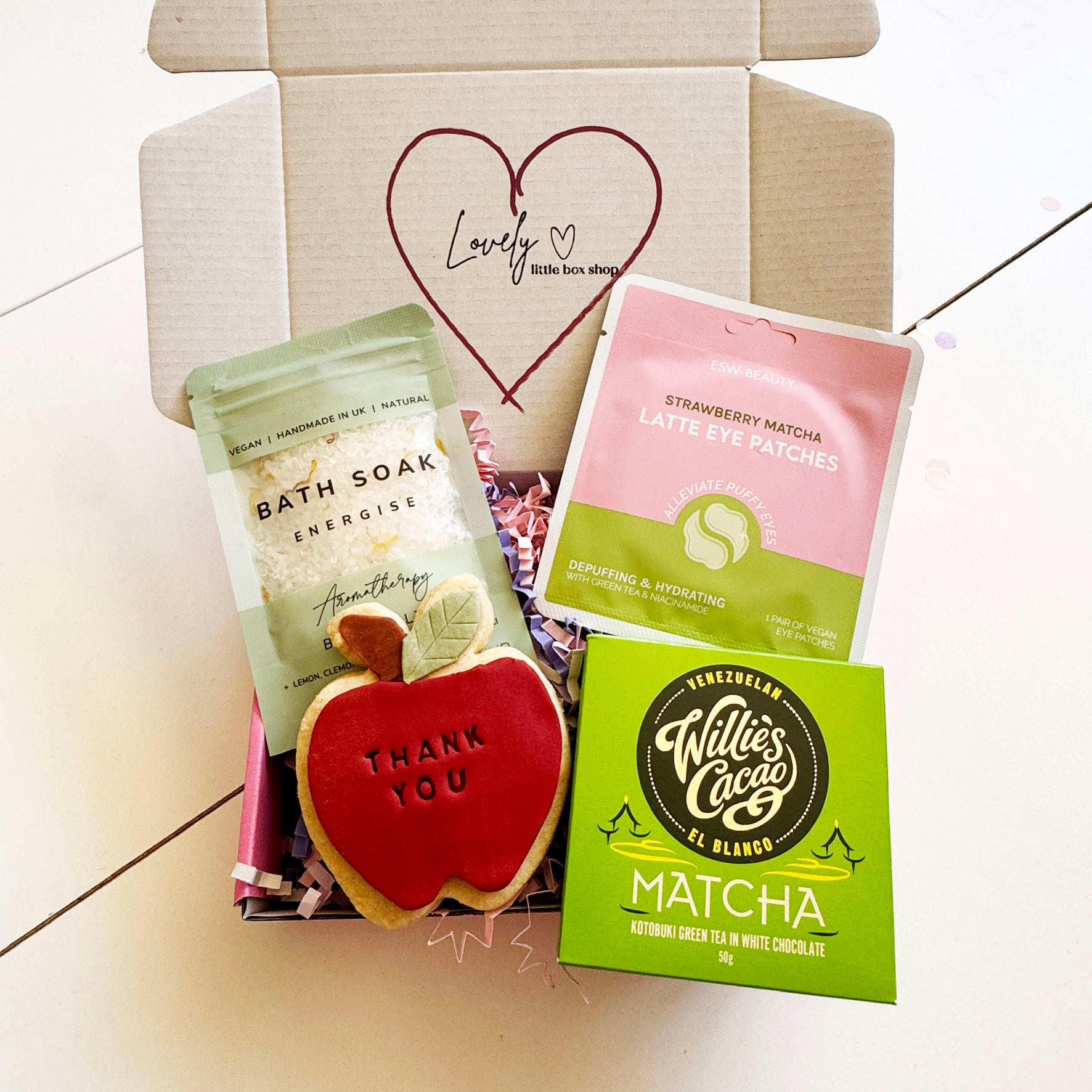 Open gift box from Lovely Little Box Shop containing a thank you apple-shaped biscuit, energising bath soak, strawberry matcha latte eye patches, and a green box of Willie’s Cacao matcha chocolate, styled on a white background.