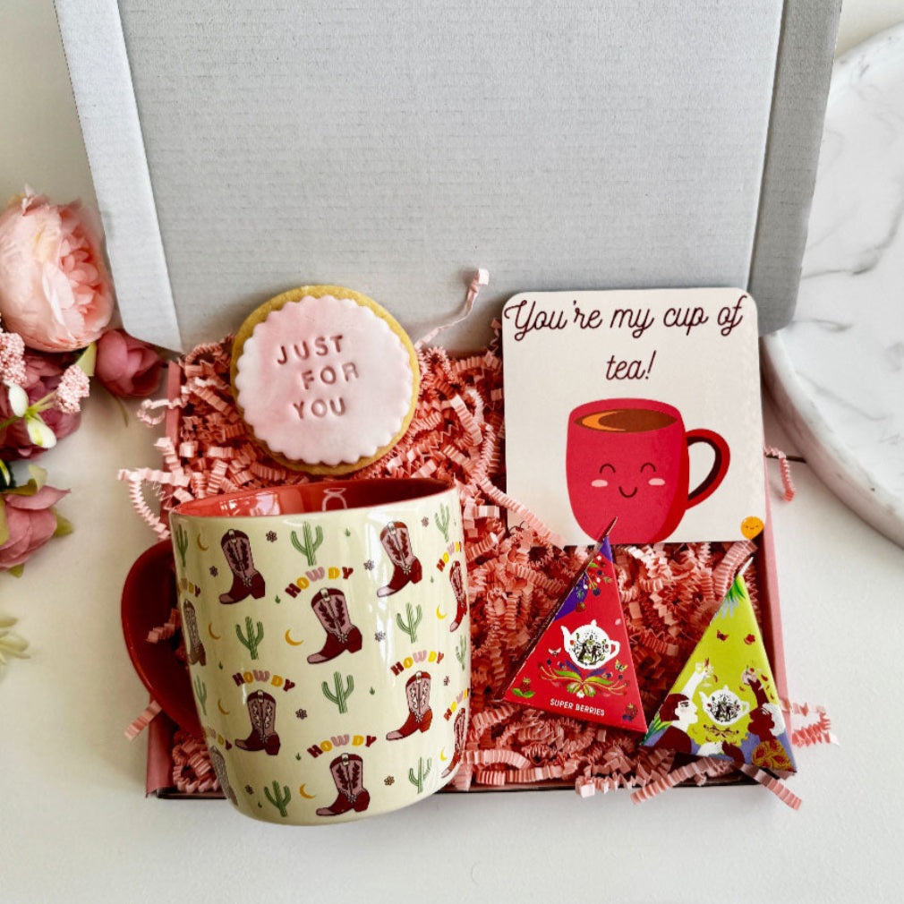 gift box containing a lavender-scented self-heating eye mask, pink tealight candle, four heart-shaped wax melts, and a rainbow enamel pin reading “Amazing Teacher,” all displayed on shredded kraft paper in a gift box.
