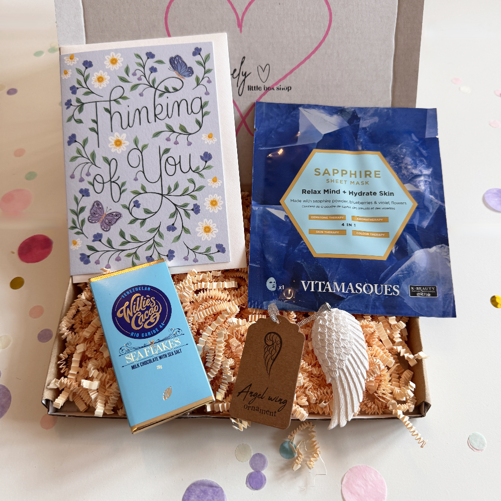 Gift box with 'Thinking of You' card, chocolate bar, Vitamasques product, and shell on a decorative background.