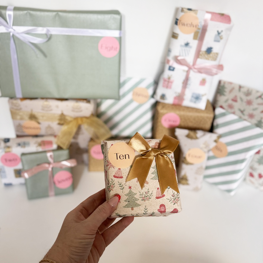 Hand holding a small gift box with a gold bow in front of other wrapped gifts.