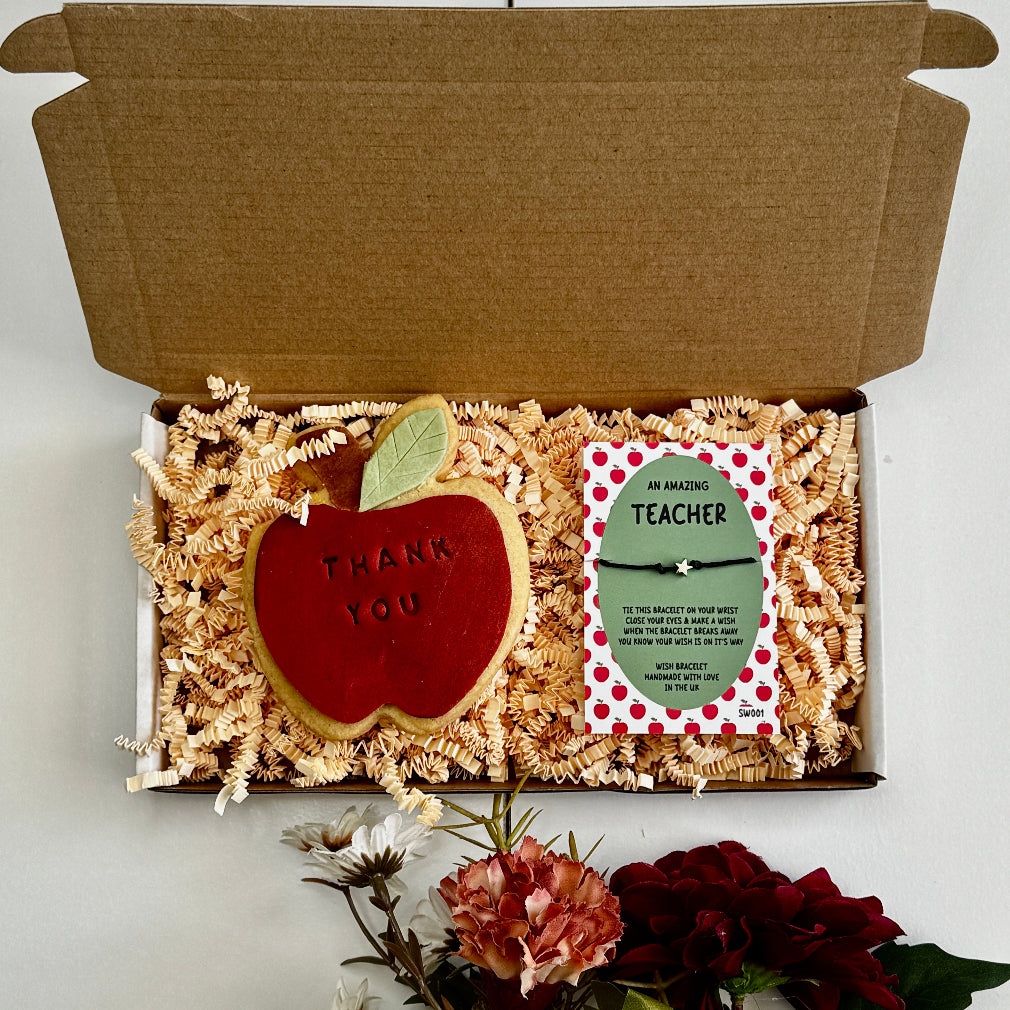 A teacher appreciation gift box featuring an apple-shaped iced biscuit with “Thank You” text and a green wish bracelet card on shredded paper filler, surrounded by flowers.
