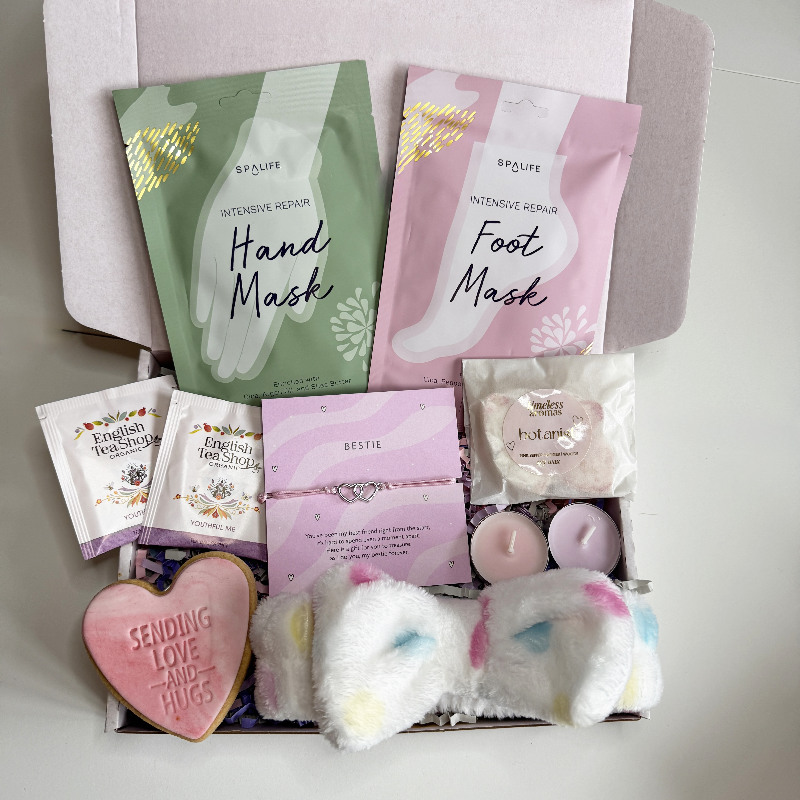 Open pastel gift box containing hand and foot masks, fluffy spa headband, heart-shaped “Sending Love and Hugs” biscuit, tea sachets, wax melt, pink tealight candles and a best friend bracelet card.