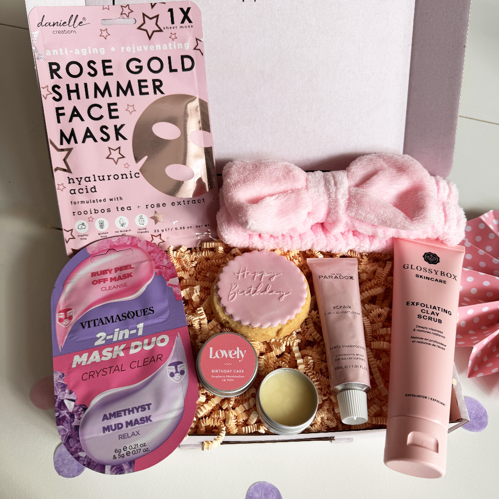 Birthday pamper gift box featuring a rose gold shimmer face mask, 2-in-1 mud and peel-off mask, exfoliating clay scrub, satin pink bow headband, lip balm, birthday biscuit, and mini skincare items, packed with colourful shredded paper.