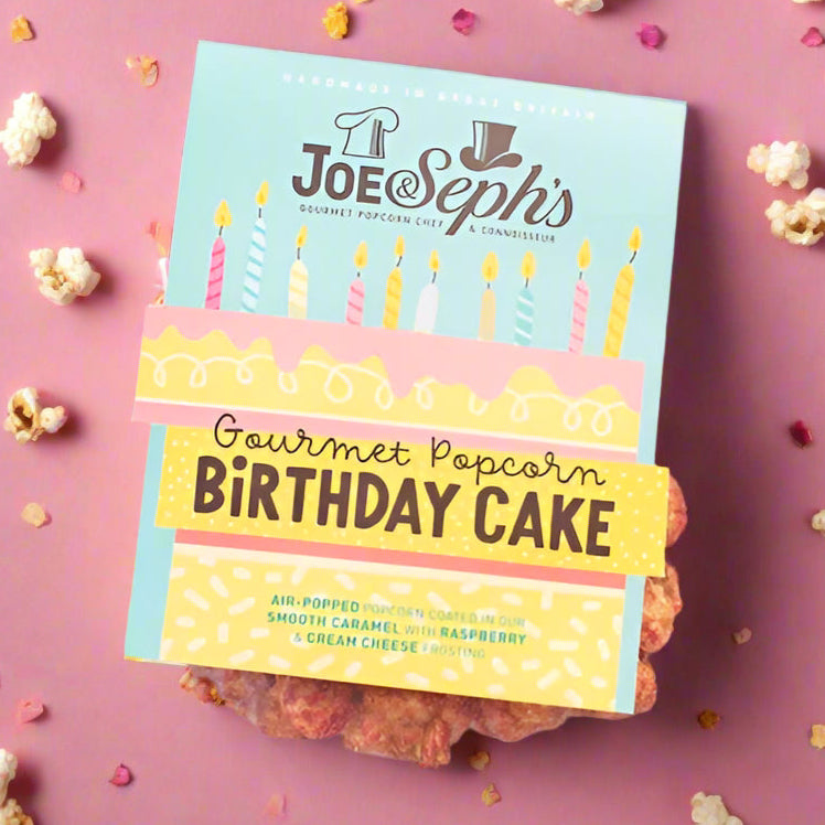 Packaged gourmet popcorn with 'Birthday Cake' flavor from Joe & Seph's, surrounded by birthday-themed decorations on a light background.