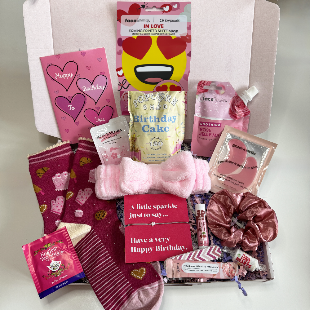 Open birthday pamper gift box with pink themed self care treats including heart socks, beauty masks, scrunchie, spa headband, birthday cake popcorn, tea sachet, lip balm and a pink happy birthday card arranged inside a gift box.