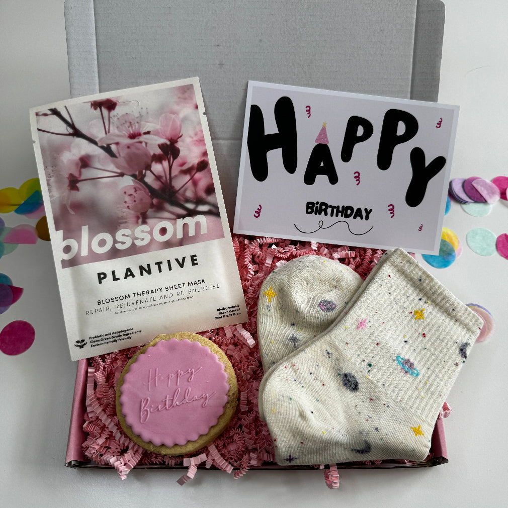Birthday-themed letterbox gift box including a pink iced “Happy Birthday” biscuit, blossom sheet mask, cute planet-themed socks, and a birthday card.
