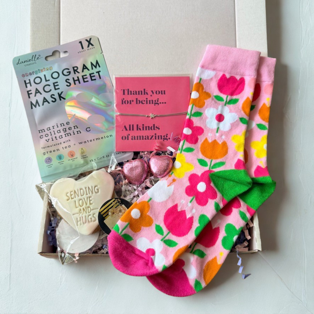A curated gift box containing a pair of pink crew socks with a colorful tulip print and green bow, a package of Danelle's hologram face mask, a heart-shaped cookie, and a pink thank you card.