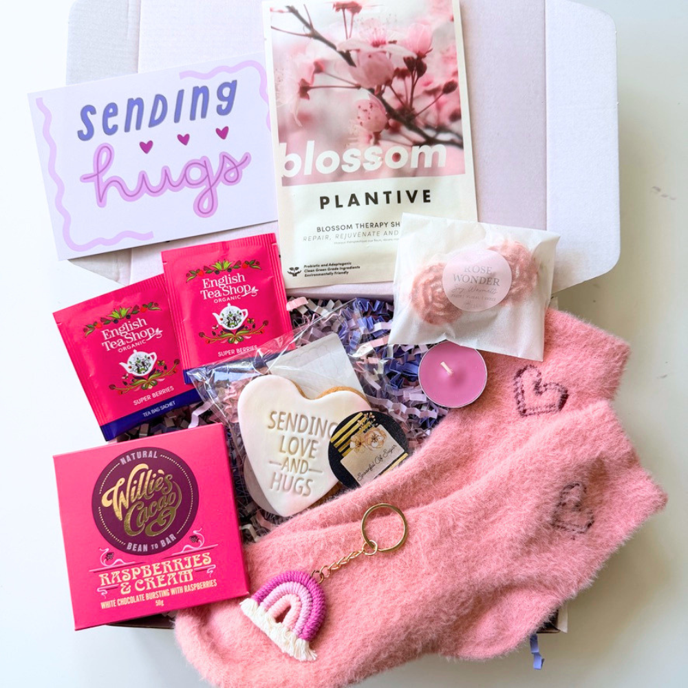 A curated gift box flat-lay featuring  pink fuzzy socks, a small pink candle, a heart-shaped cookie, a pink and white keychain, two pink tea packets, a card reading 'Sending hugs,' and a face mask 'blossom PLANTIVE' on a white background.