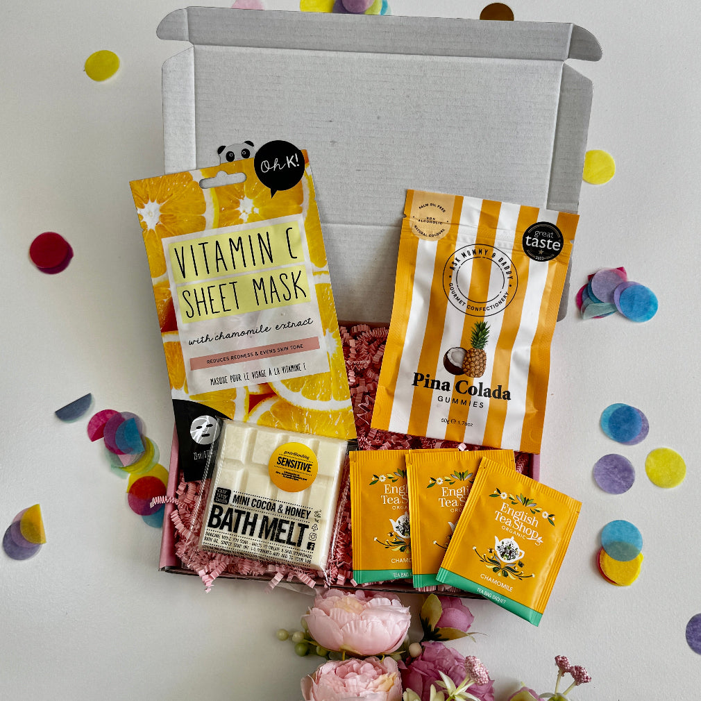 A letterbox gift set featuring a vitamin C face mask, Pina Colada gummies, mini cocoa & honey bath melt, and chamomile tea sachets, styled with pastel confetti and flowers.