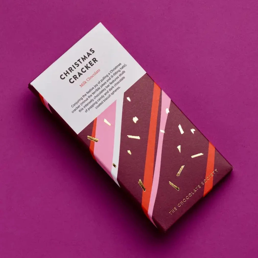 A boxed chocolate bar package labeled 'Christmas Cracker' by The Chocolate Society, photographed on a solid purple background. The box is dark burgundy with pink and orange diagonal stripes, white text, and small gold confetti accents.