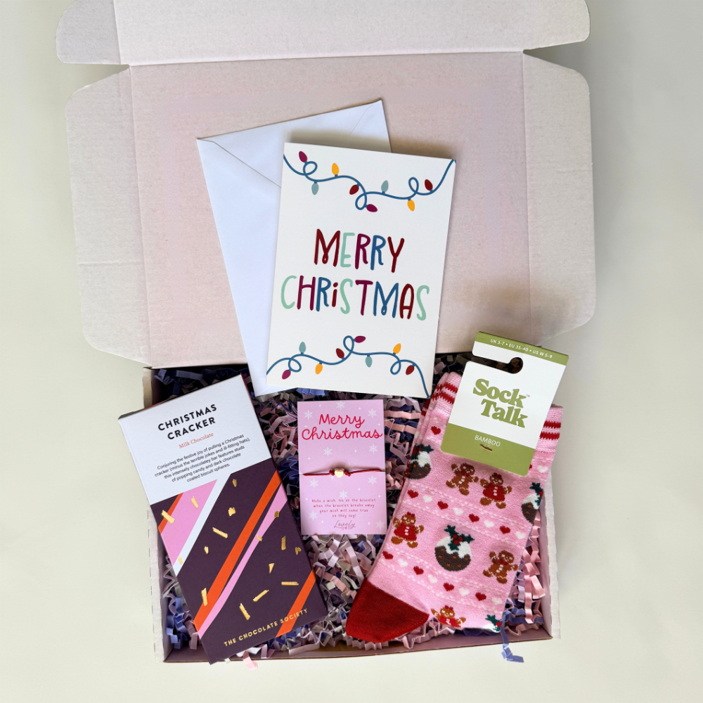 A curated Christmas gift box set displayed from above inside an open kraft paper box. The set includes a white Christmas greeting card with colorful string lights and the words 'MERRY CHRISTMAS,' a white envelope, a pink and white patterned pair of socks with a 'Sock Talk' tag, a chocolate bar labeled 'The Chocolate Company,' and a small pink card reading 'Merry Christmas.