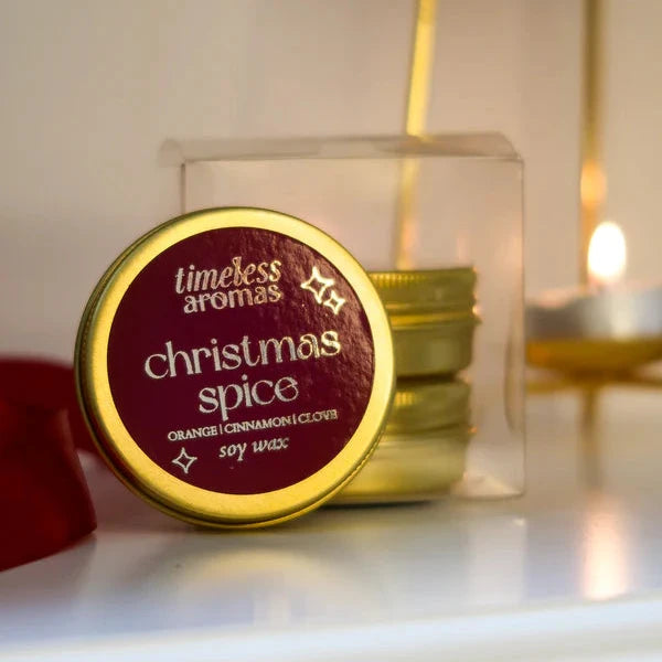 Candle labeled 'christmas spice' by 'timeless aromas' on a white surface with a blurred candle in the background.