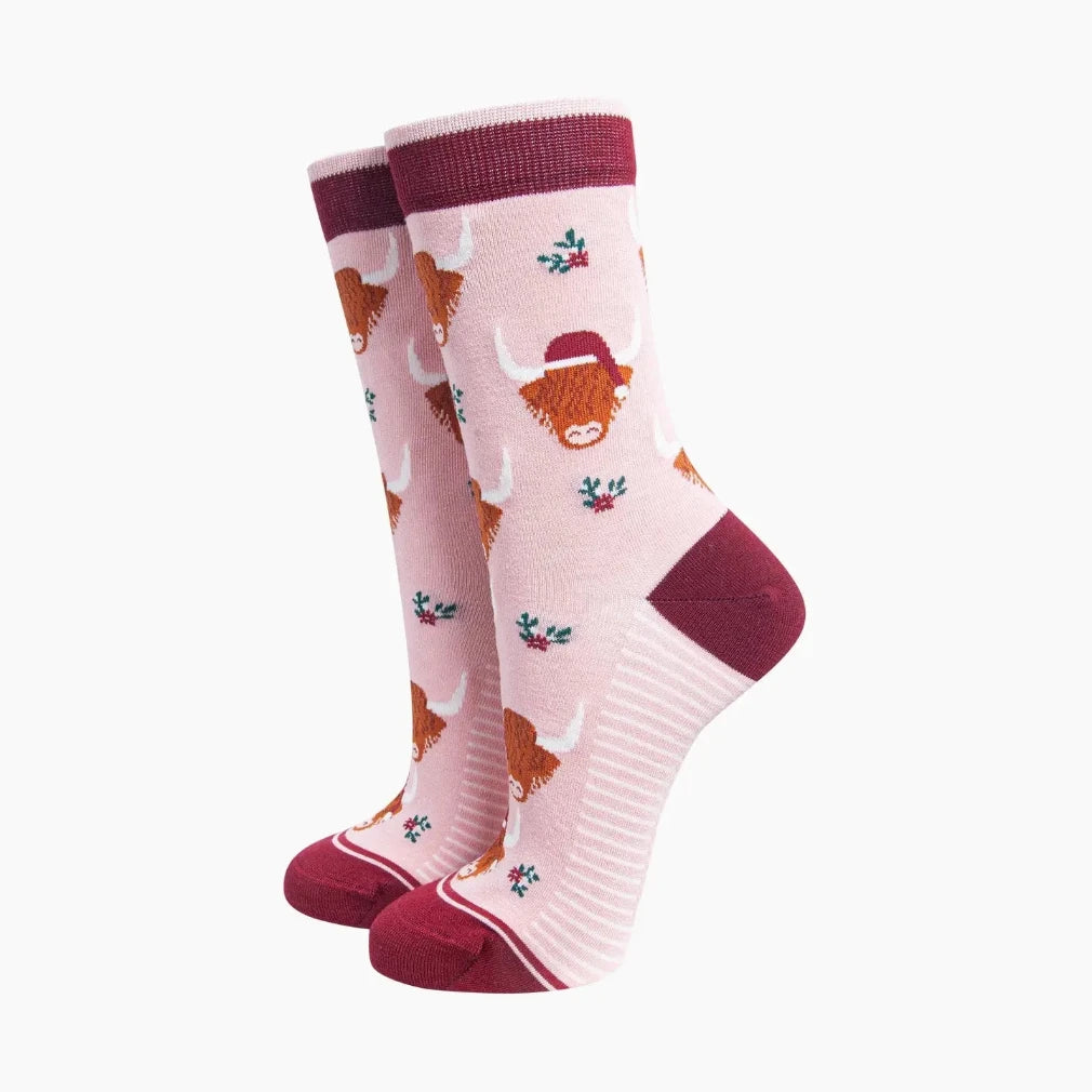 A pair of pink crew socks featuring a pattern of orange Highland cows, small green holly leaves, and red cuffs and heels, displayed against a plain white background.
