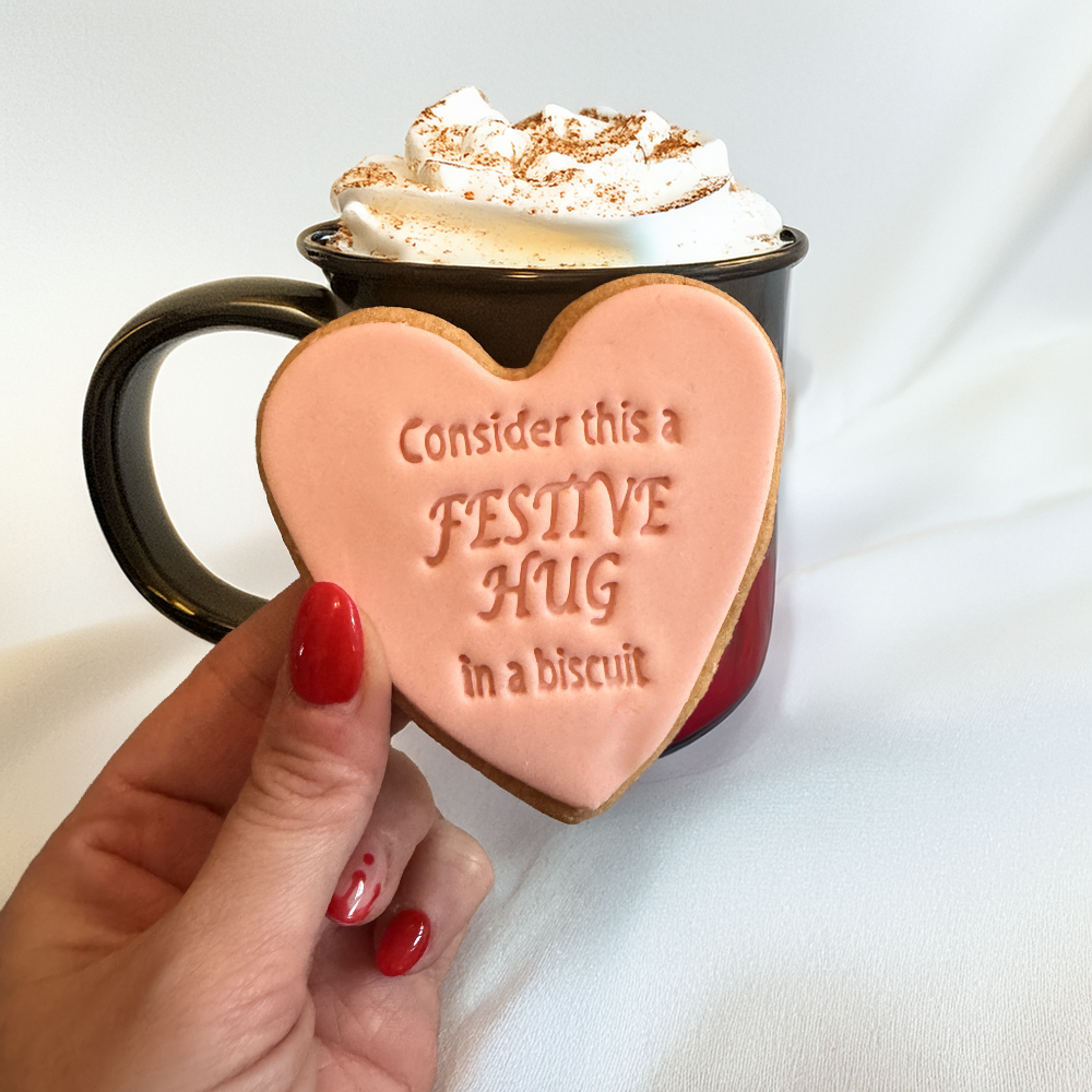 Heart-shaped cookie with text held in front of a mug of hot chocolate.