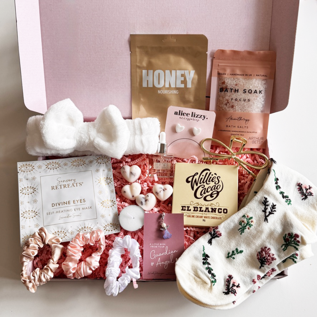 Gifting box with various products including a bath soak, soap, and skincare items on a light pink background.