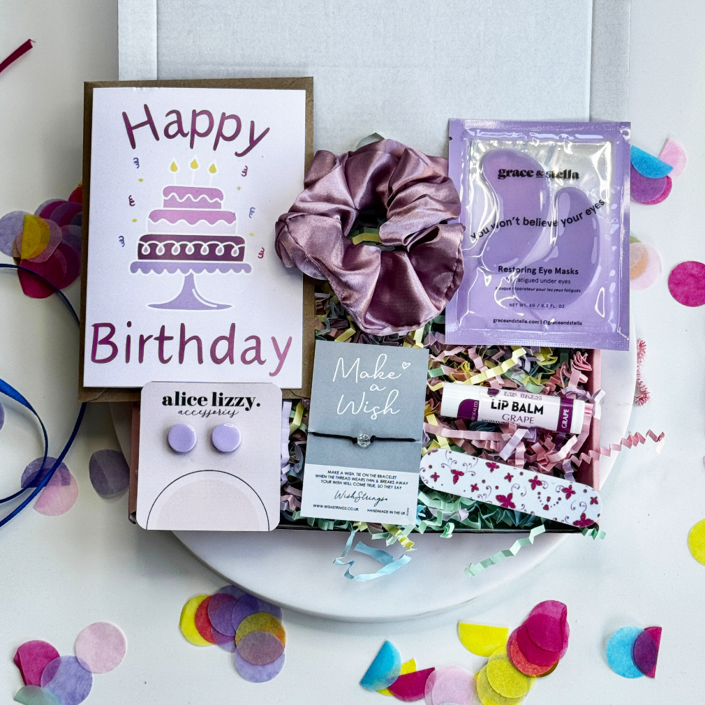 Birthday gift box for her with “Happy Birthday” card, scrunchie, earrings, wish bracelet, grape lip balm, eye mask, and nail file on pastel shredded paper.