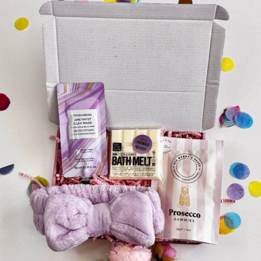A flat-lay image of a pastel-themed pamper gift box including a clay mask, lavender bath melt, prosecco-flavoured sweets, and a soft lilac headband, surrounded by confetti and pink flowers.
