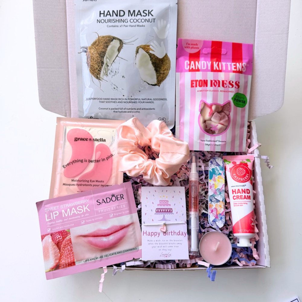 A curated gift box filled with beauty and snack items, including a hand mask pouch, a pink striped candy kitten bag, a pink scrunchie, a lip mask packet, a hand cream tube, a lip gloss tube, and a small candle, arranged in crinkle paper inside an open box.