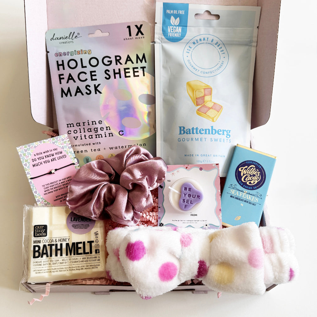 Assorted beauty and lifestyle products including a hologram face sheet mask, bath melt, scrunchie, and chocolate bar on a white surface.