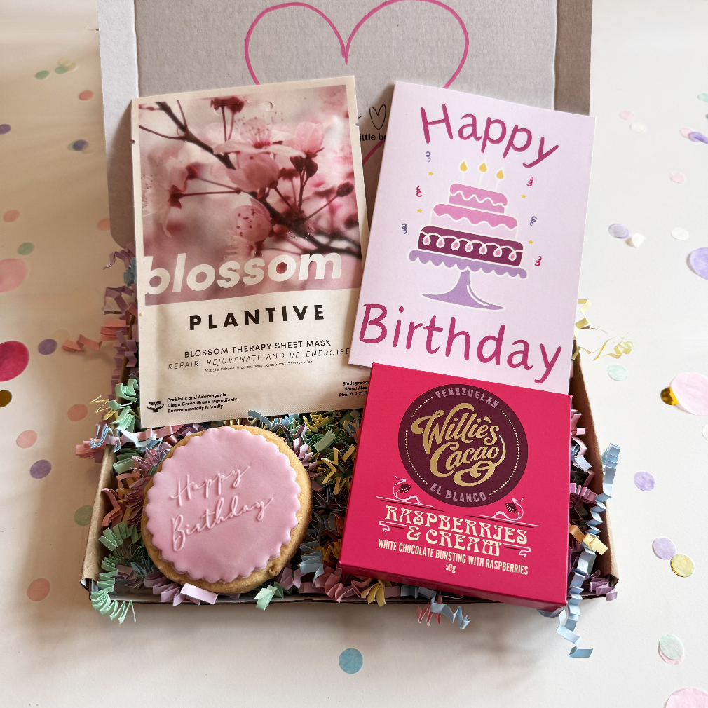 Gift set with 'blossom' plantative mask, 'Happy Birthday' card, and 'Willie's Cacao' chocolate on a decorative background.