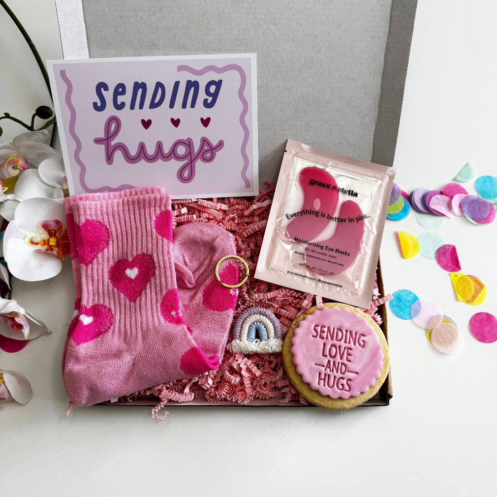 Teen pamper gift box with heart-print socks, pink under-eye masks, a “Sending Love and Hugs” iced biscuit, and rainbow keyring, all arranged in pink shredded paper with confett