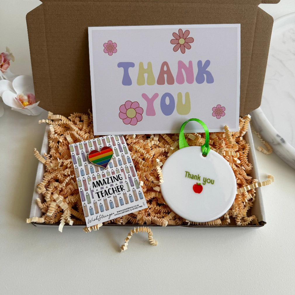 Teacher thank-you gift box including a pastel "THANK YOU" card, rainbow heart pin badge on “Amazing Teacher” backing card, and white ceramic hanging ornament with “Thank you” and red apple design, all packed in shredded kraft paper.