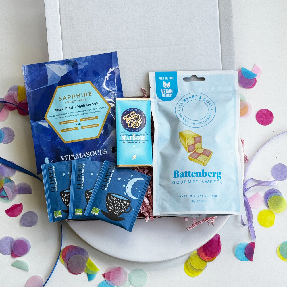 Relaxation gift box with sapphire face mask, Pukka herbal teas, vegan Battenberg sweets, and Willie’s Cacao sea salt chocolate on pink shredded paper.