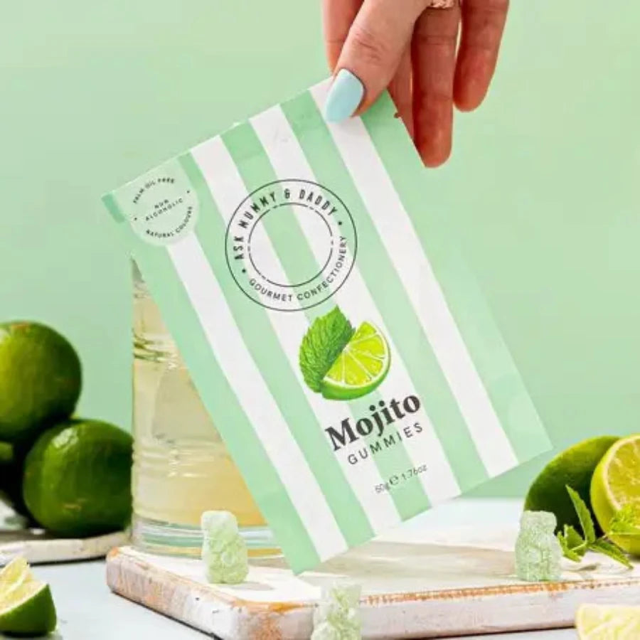 Hand holding a green and white package of Mojito Gummies with limes and a glass in the background.