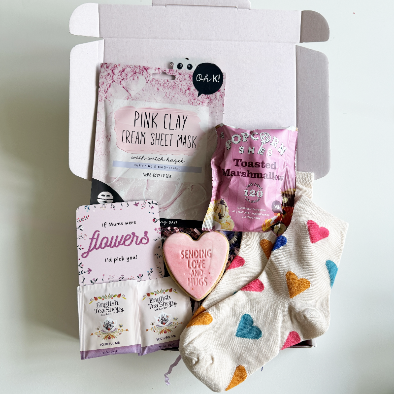 Open pastel gift box filled with heart-patterned socks, a pink clay face mask, toasted marshmallow popcorn, tea sachets and a pink heart biscuit that says “Sending Love and Hugs,” alongside a floral Mother’s Day card.
