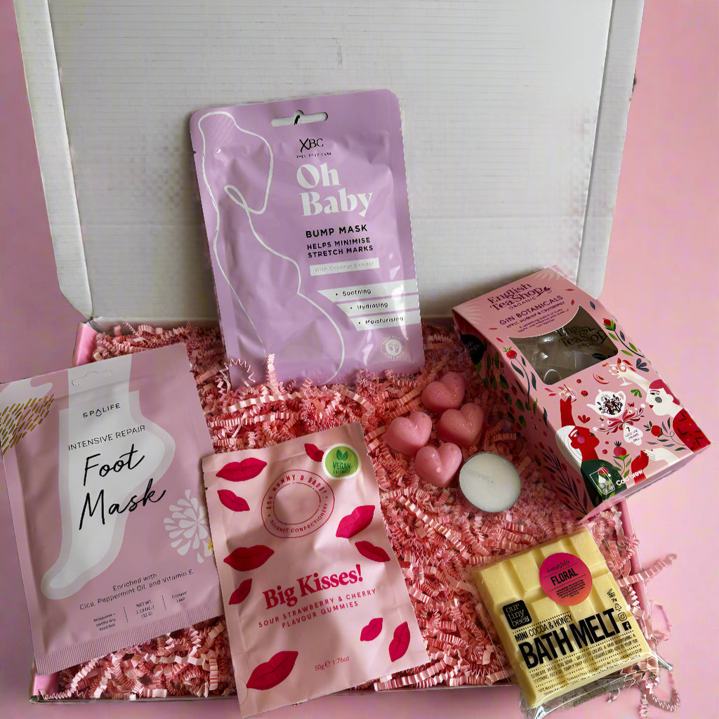 Pregnancy gift box with pampering and treat items including a bump mask, foot mask, bath melt, tea, heart-shaped melts, a tealight candle, and vegan sweets.