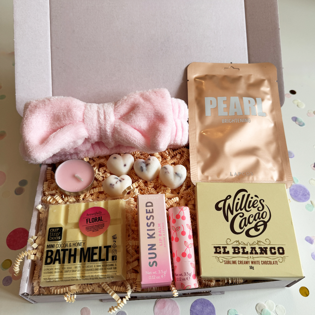 Gifting box with bath products including a pink bow, bath melt, and chocolate bar on a colorful background.