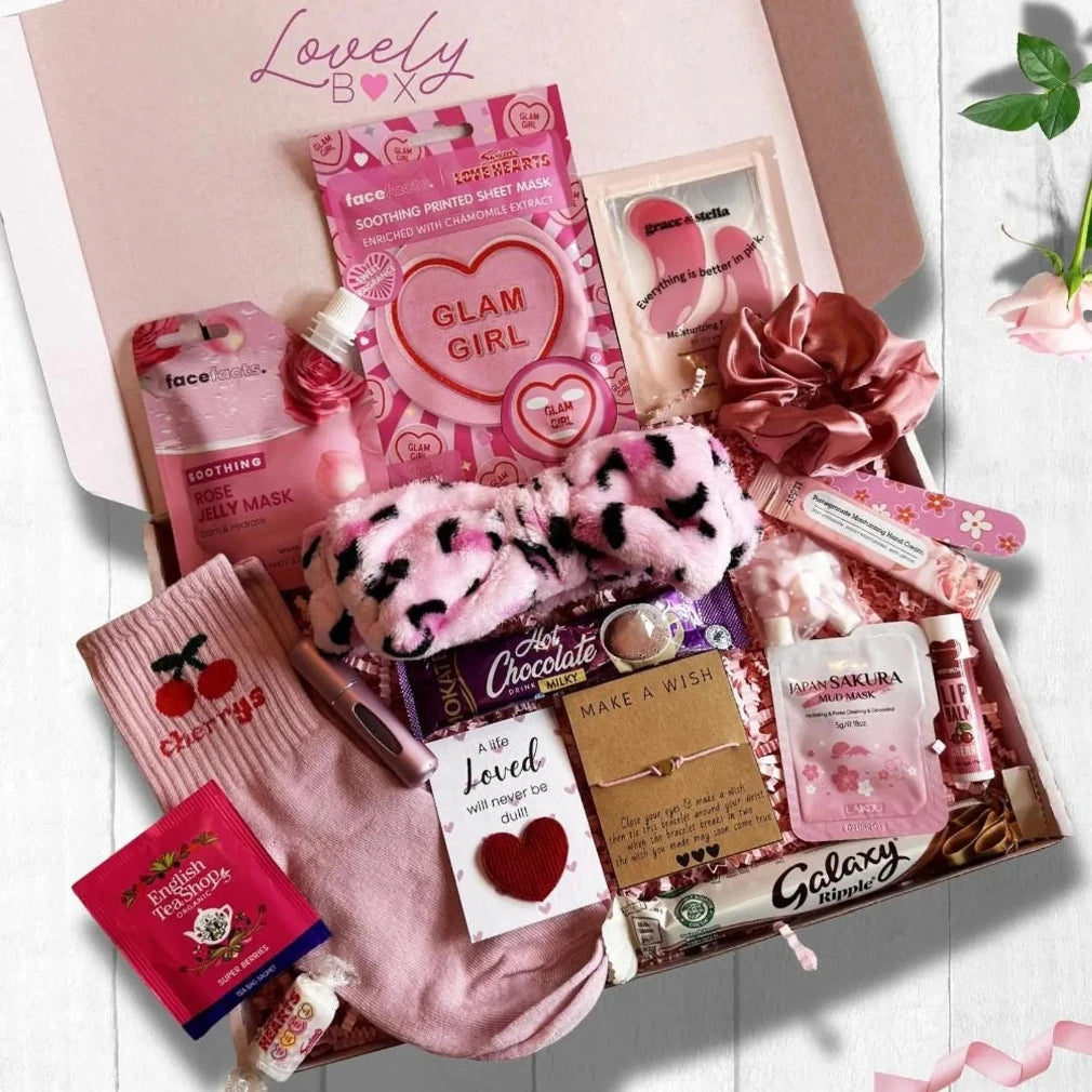 A birthday themed gift box filled with pink beauty and self-care items, including a pink fuzzy headband, cherry-print socks, a pink heart mask, a chocolate bar, and lip balm, arranged in a box labeled 'Lovely Box'.