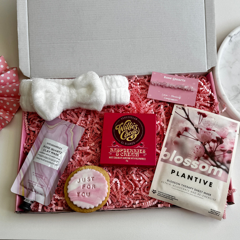 Flat lay of a pink pamper gift box including a white bow headband, raspberry chocolate, rose quartz clay mask, blossom sheet mask, rose quartz crystal, and iced biscuit reading “just for you”.
