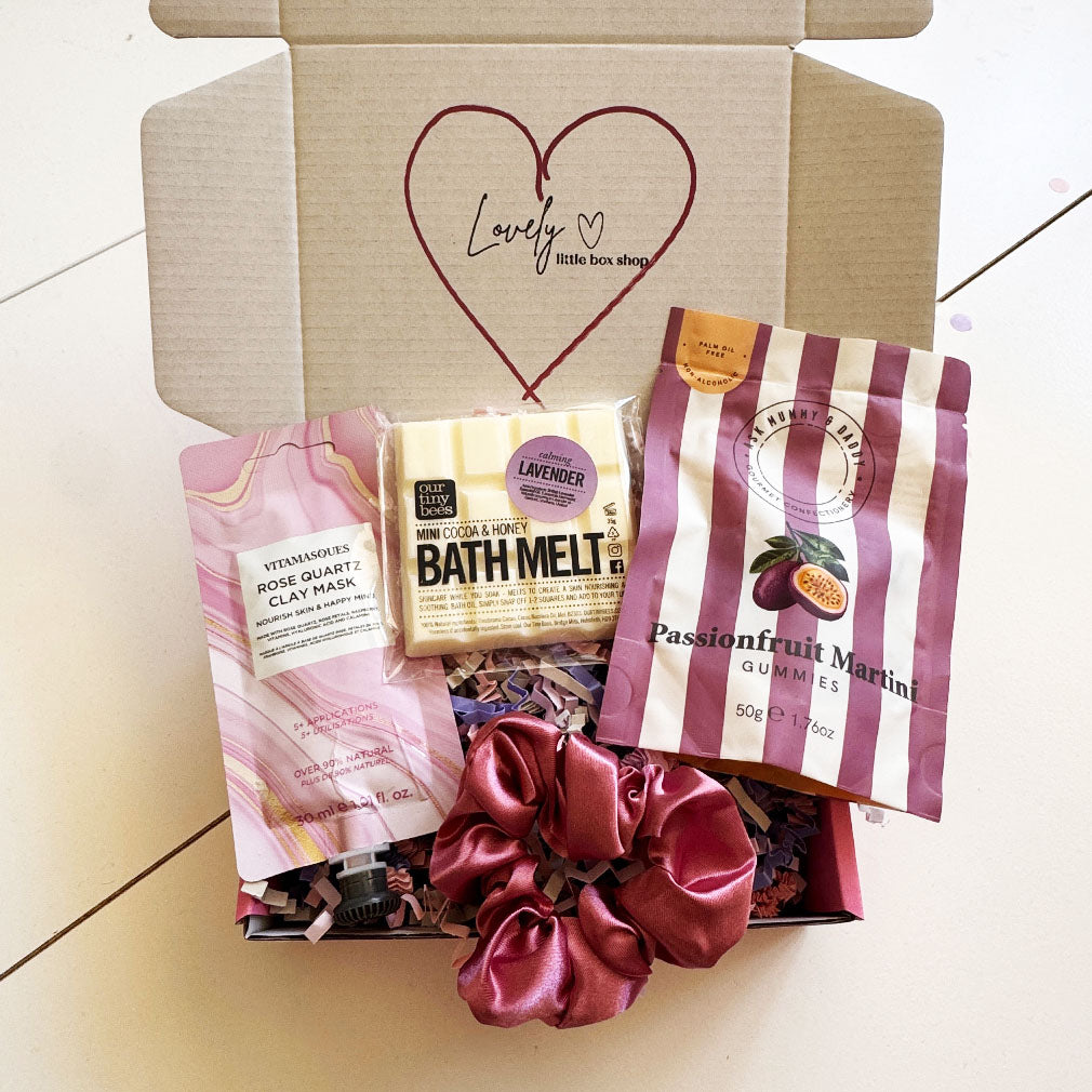 Open pink-themed gift box from Lovely Little Box Shop containing a rose quartz clay mask, lavender bath melt bar, passionfruit martini gummies in striped packaging, and a pink satin scrunchie on pink shredded filling 