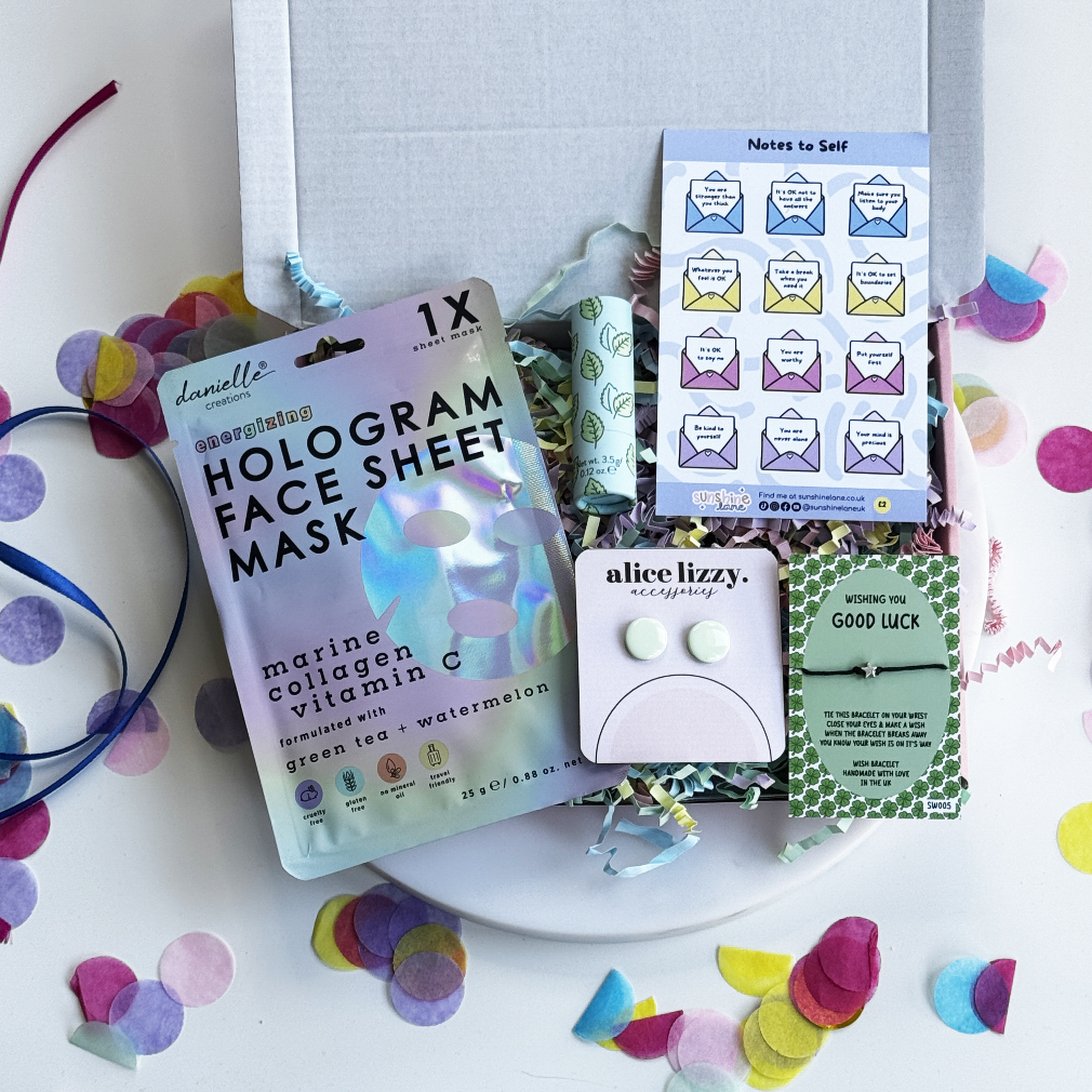 Assorted items including a hologram face sheet mask, earrings, and cards on a white surface with colorful confetti.