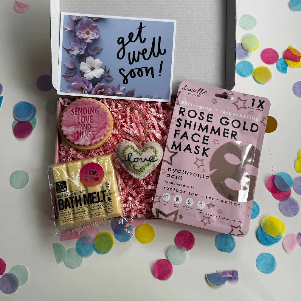 Get well soon gift box including a pink shimmer face mask, floral bath melt, “Sending Love and Hugs” iced biscuit, embroidered heart ornament, and a floral “Get Well Soon” card, surrounded by colourful confetti.