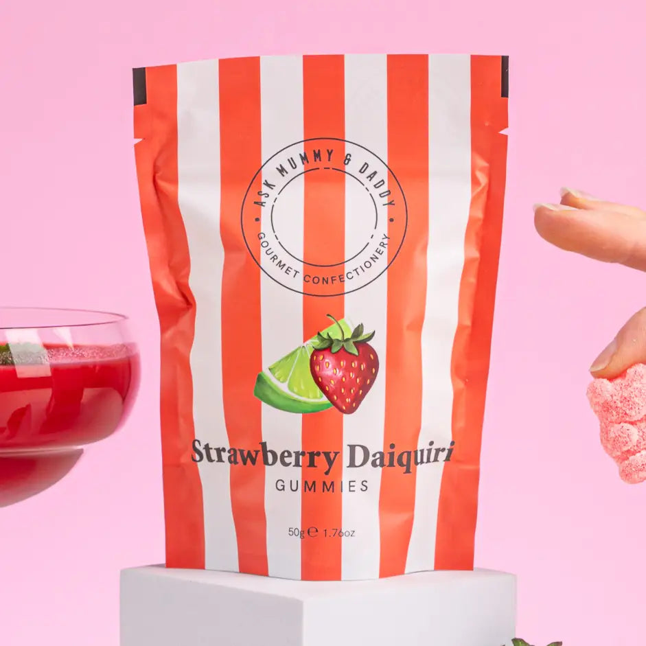 A stand-up pouch of Art Mummy & Daddy Strawberry Daiquiri Gummies, featuring red and white vertical stripes and a strawberry and lime graphic, set against a pink background with a hand holding a pink gummy candy on the right.