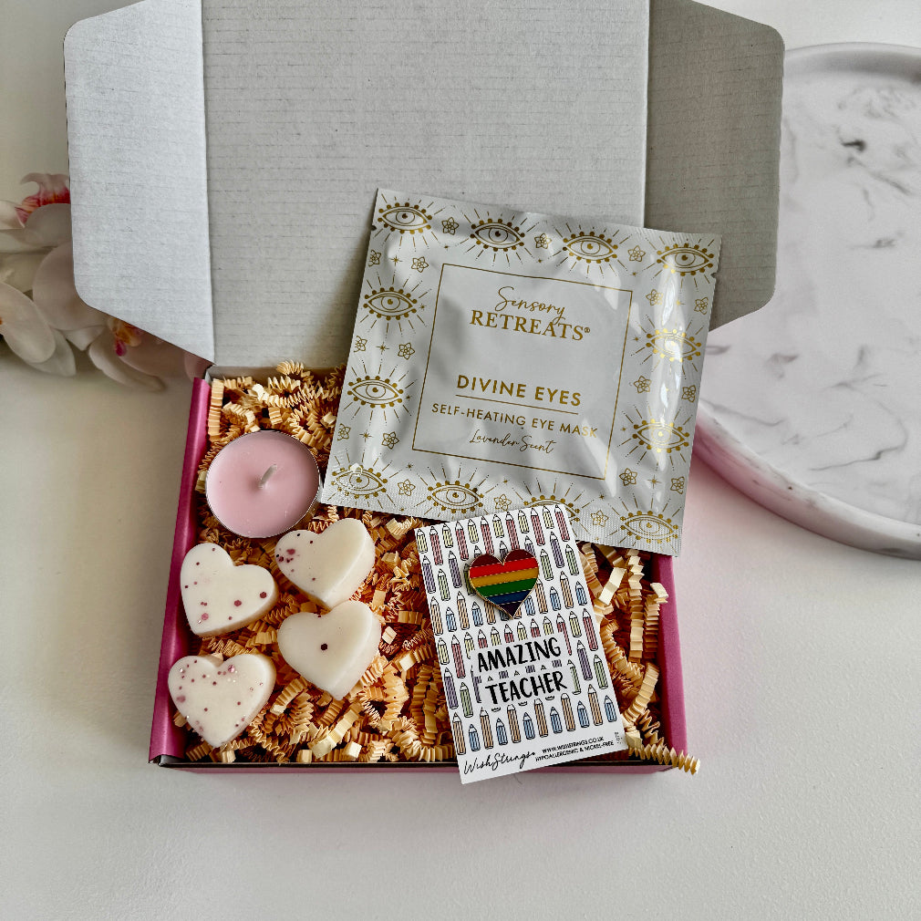 Teacher thank-you gift box containing a lavender-scented self-heating eye mask, pink tealight candle, four heart-shaped wax melts, and a rainbow enamel pin reading “Amazing Teacher,” all displayed on shredded kraft paper in a gift box.