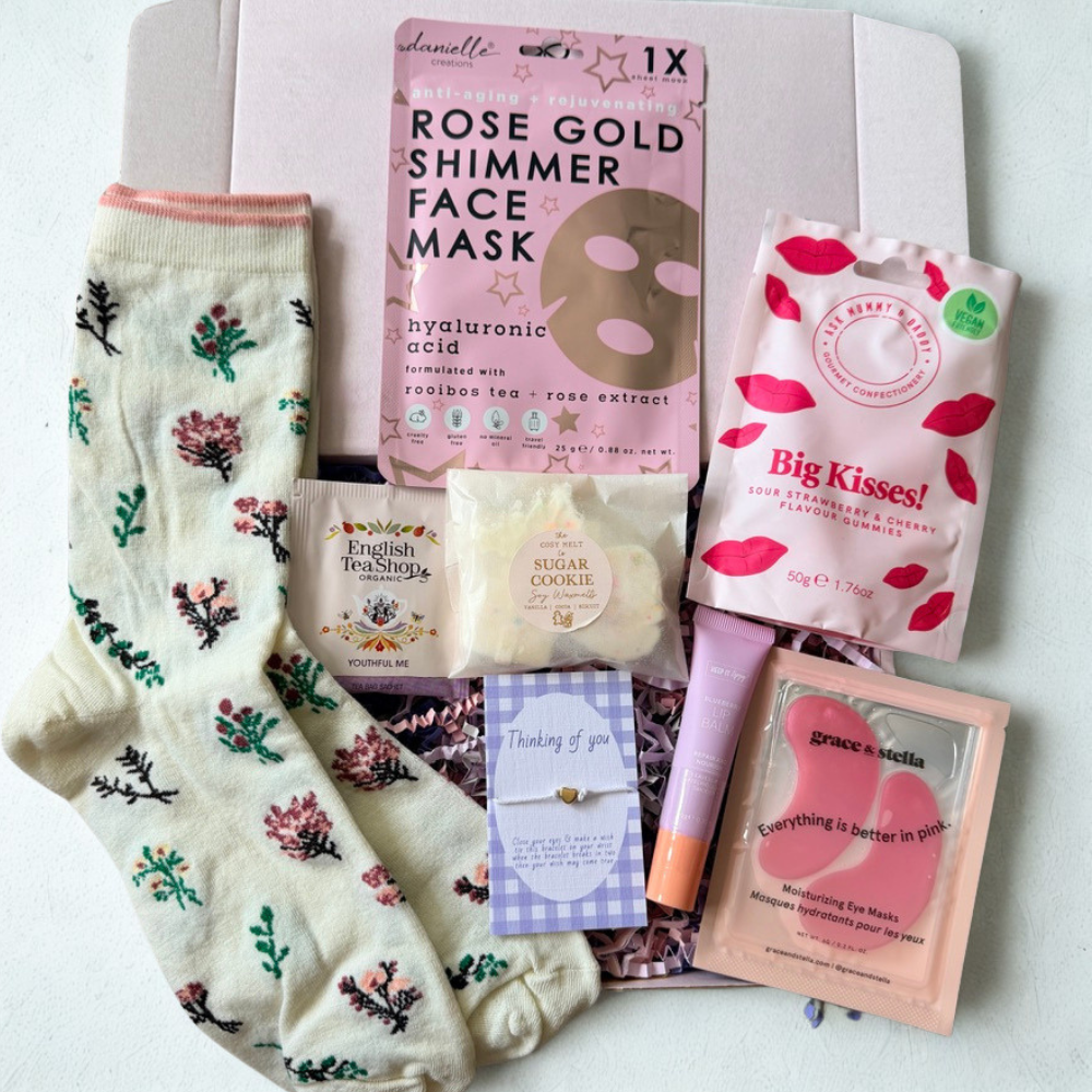 A curated gift set arranged in a flat lay on a white background, featuring a pair of cream socks with a floral print, a pink rose gold face mask packet, a pink lip treatment tube, a sugar cookie bar, a tea shop packet, and other small beauty and gift items.
