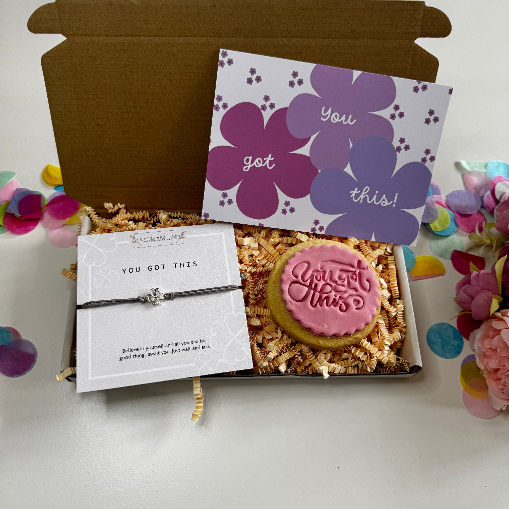 Flat lay of a letterbox gift including a “You Got This” iced biscuit, motivational bracelet with a silver charm, and a floral encouragement postcard.