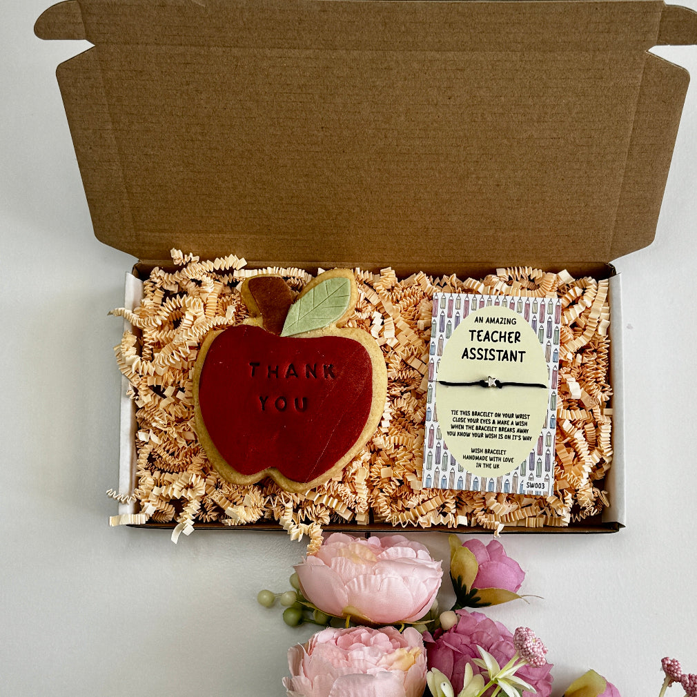 A teacher appreciation gift box featuring an apple-shaped iced biscuit with “Thank You” text and a pencil patterned wish bracelet card on shredded paper filler, surrounded by flowers.