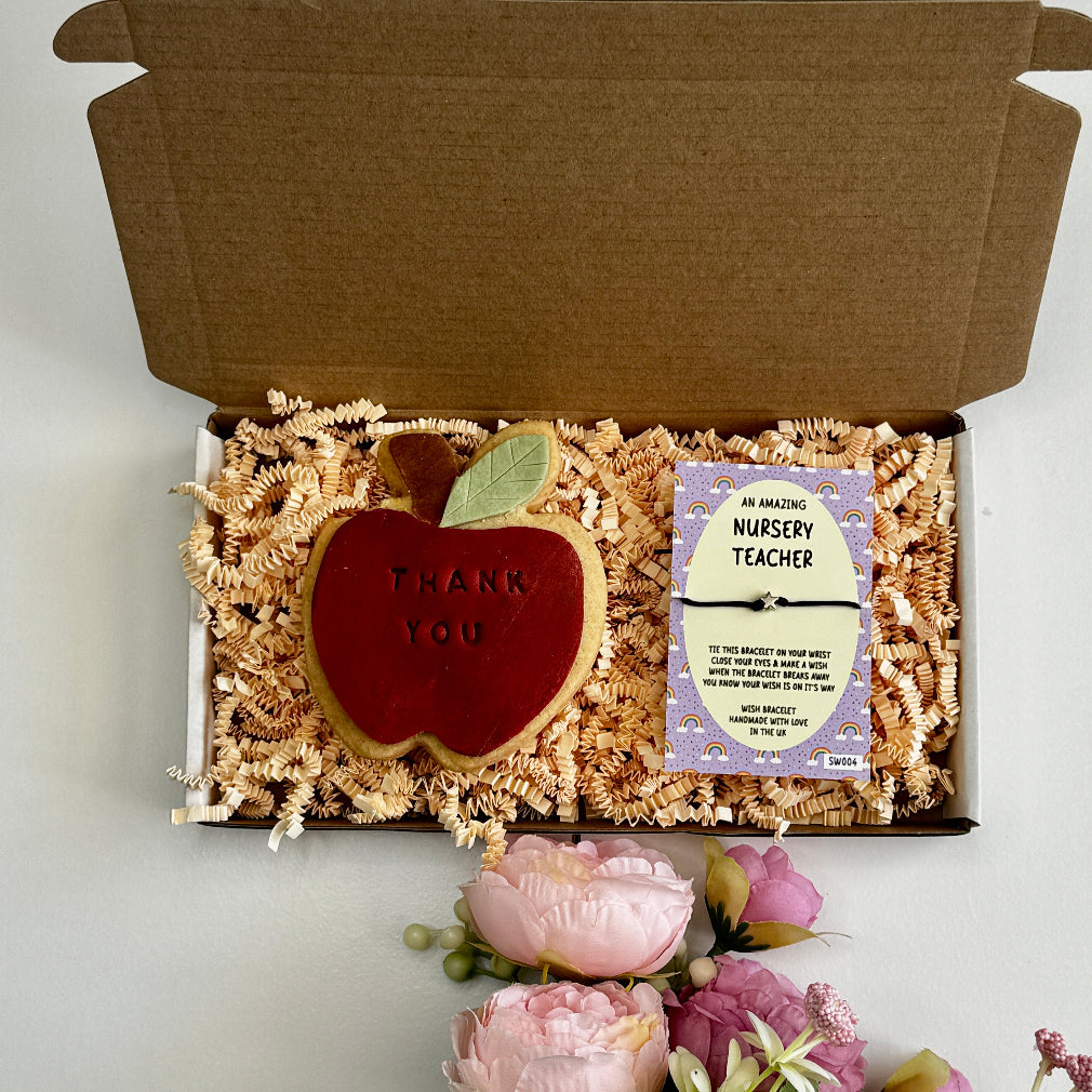 A teacher appreciation gift box featuring an apple-shaped iced biscuit with “Thank You” text and a green wish bracelet card on shredded paper filler, surrounded by flowers.