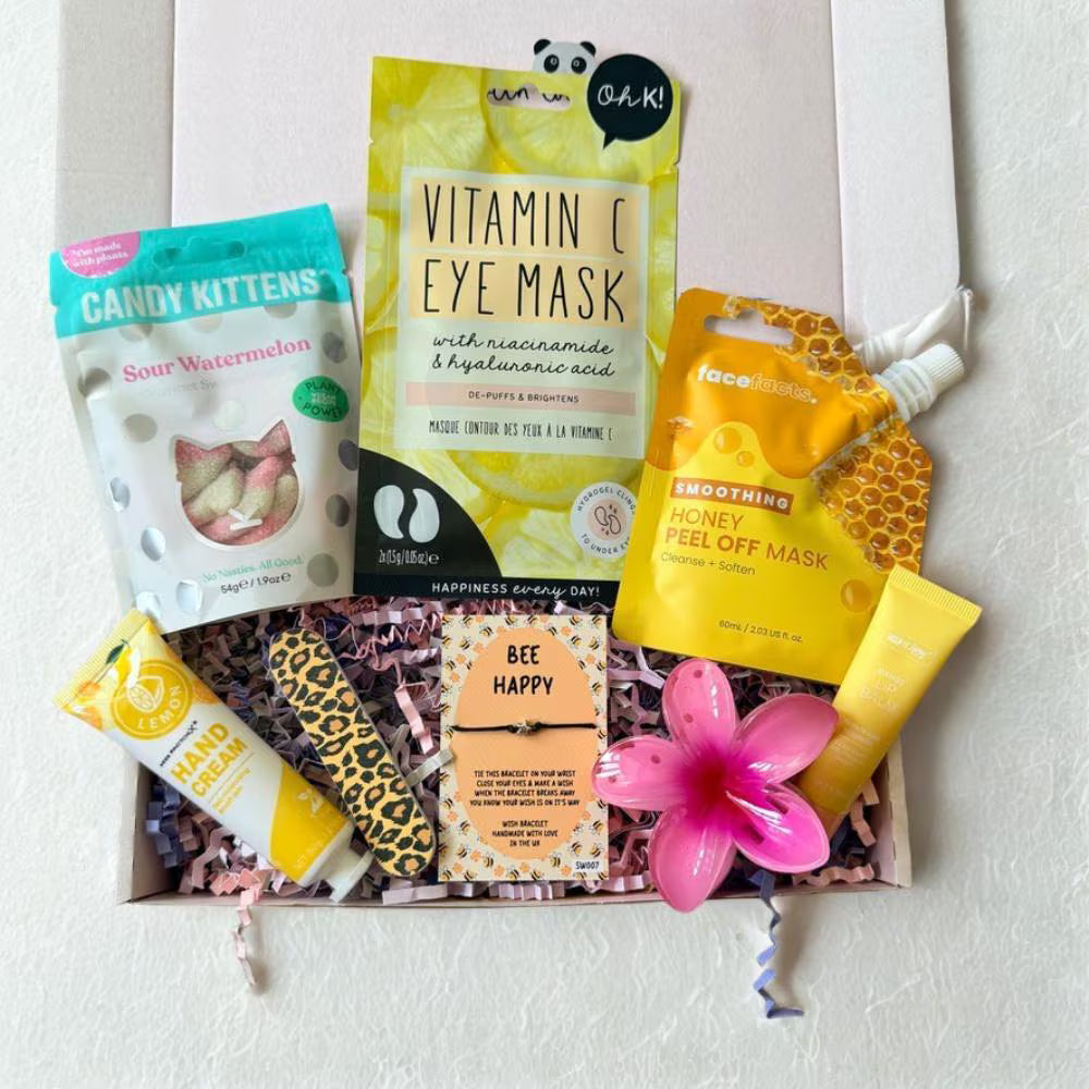 An open gift box filled with skincare and beauty products, including a Vitamin C Eye Mask, Honey Feel Off Mask, a tube of hand cream, a leopard-print lip balm, a small card reading 'BEE HAPPY,' and a pink flower, arranged on a white background.