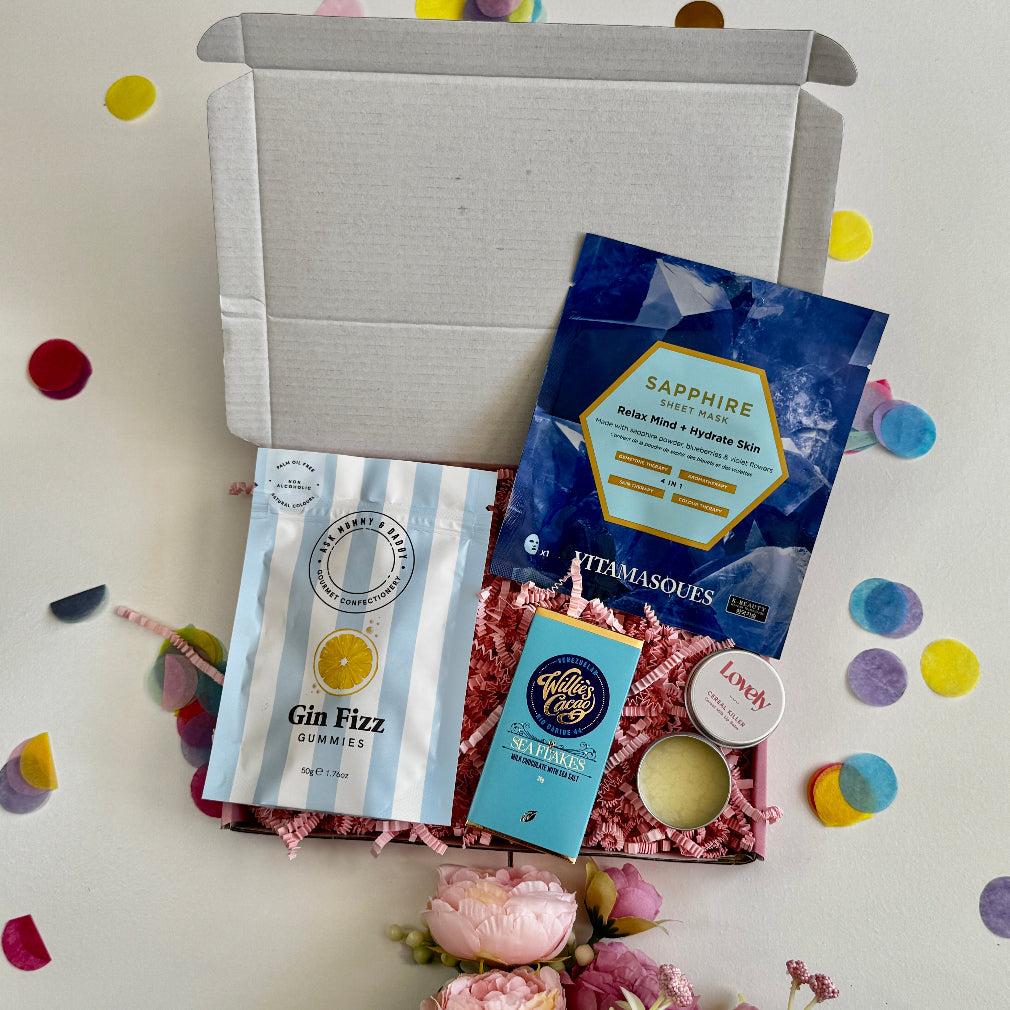A letterbox gift set including a sapphire face mask, gin fizz gummies, Willie's Cacao chocolate, lip balm, and a candle, styled with pink shredded paper and confetti.