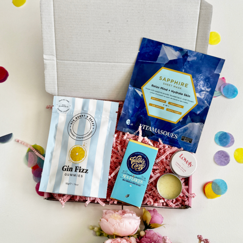A letterbox gift set including a sapphire face mask, gin fizz gummies, Willie's Cacao chocolate, lip balm, and a candle, styled with pink shredded paper and confetti.